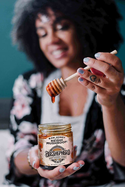woman holding honey dripper to camera above jar of reishi mushroom honey with heart tattoo on finger